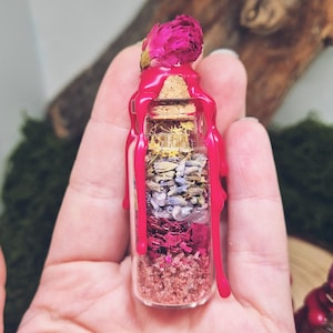 Könnte beinhalten: Eine kleine Glasflasche gefüllt mit Schichten aus getrockneten Blumen, Kräutern und Kristallen, verschlossen mit einem Korken und leuchtend rosa Wachs. Oben auf der Flasche sitzt eine getrocknete rosa Rose. Die Flasche wird in einer Hand gehalten.
