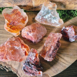 Könnte beinhalten: Eine Sammlung roher, grob geschnittener Kristalle in Orange-, Rot- und Klartönen. Die Kristalle variieren in Größe und Form, mit sichtbaren inneren Mustern und Texturen. Sie sind auf einer Holzoberfläche angeordnet.