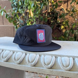 May include: A black baseball cap featuring a rectangular patch on the front. The patch has a blue border and a red background with the text "BUFFALO FOOTBALL" in blue letters, accompanied by a white line-art illustration of a buffalo head.
