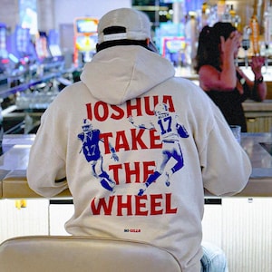 May include: A cream-colored hoodie with a graphic print of two football players and the text "Joshua Take the Wheel" in red and blue. The text "Go Bills" is printed in small letters below the graphic.