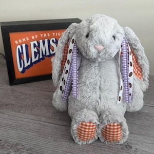 May include: A plush gray bunny with long ears and a pink nose. The bunny has patterned ribbons and orange and white checkered accents on its ears and feet. In the background is a framed sign with the text "CLEMS" and "HOME OF THE TIGERS".