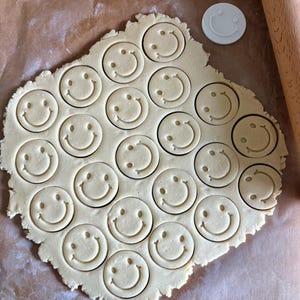 Op de afbeelding: Een uitgerolde lap lichtbeige koekjesdeeg met meerdere cirkelvormige smiley-afdrukken in het oppervlak. Een enkele witte ronde plastic koekjesvorm met een smiley-ontwerp en een houten deegroller zijn zichtbaar in de afbeelding.