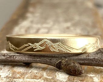 May include: A gold band ring with a mountain range design engraved on the top. The ring is sitting on a branch.