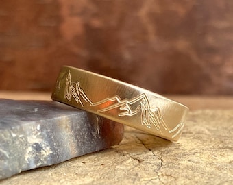 May include: A gold band with a mountain range design engraved on the top. The band is sitting on a piece of rough, brown rock.