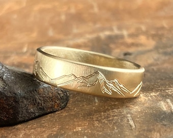May include: A gold wedding band with a mountain range design engraved on the outside of the band.