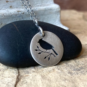 May include: A silver necklace with a round pendant featuring a cut-out silhouette of a cardinal bird perched on a branch.