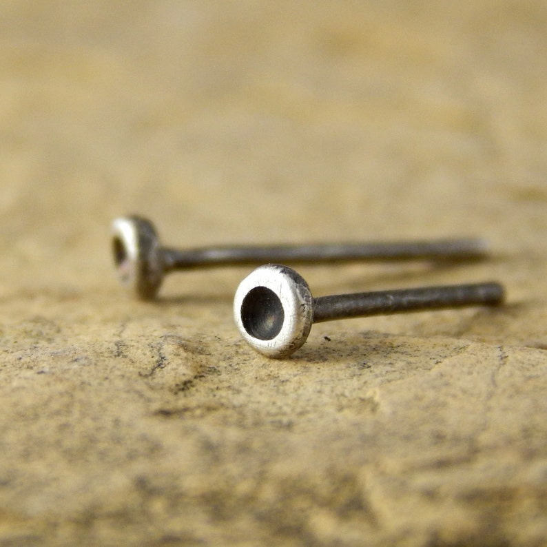 May include: Two silver stud earrings with a small, circular design. The earrings have a post and a rounded, hollowed-out top. The background is a textured, tan surface. The earrings are shown in a close-up shot.