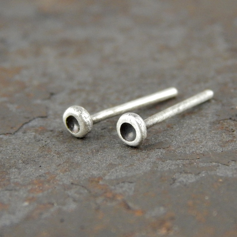 May include: A pair of small, silver stud earrings. Each earring features a rounded, textured head with a dark, recessed center, connected to a slender post. The earrings are displayed on a textured, gray surface.