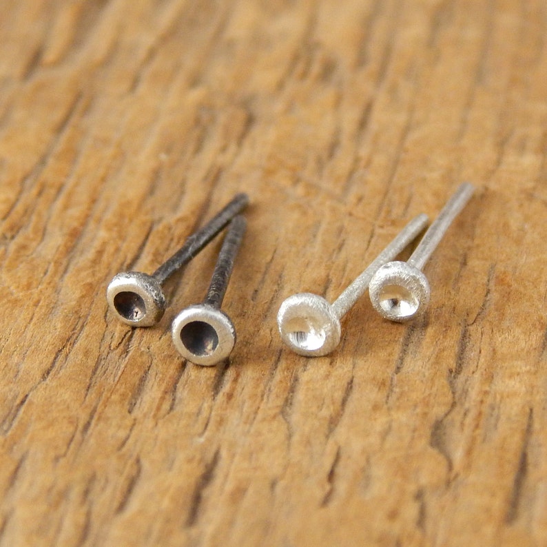 May include: Two pairs of small, silver stud earrings. One pair is a darker, oxidized silver, and the other is a brighter, polished silver. Each earring features a rounded, cup-like design on the end of a thin post.
