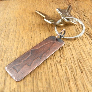 May include: A rectangular copper keychain with a fish silhouette design. The keychain is attached to a silver keyring with two keys. The copper has a rustic, aged appearance, and the background is a wooden surface.