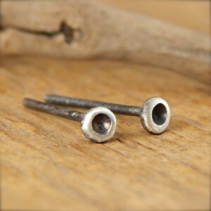 May include: Two small, silver-colored stud earrings. Each earring has a rounded, slightly textured head with a dark center, connected to a thin, straight post. The earrings are displayed on a light brown wooden surface.