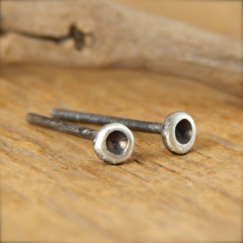 May include: Two small, silver-colored stud earrings. Each earring has a rounded, slightly textured head with a dark center, connected to a thin, straight post. The earrings are displayed on a light brown wooden surface.