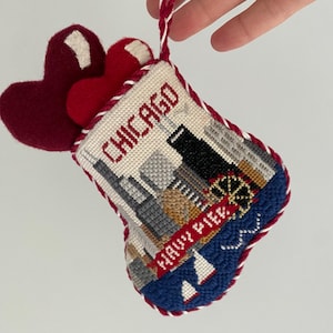 May include: A red and white stocking ornament with a stitched image of the Chicago skyline and the words "Chicago" and "Navy Pier". The stocking is filled with two red felt hearts.