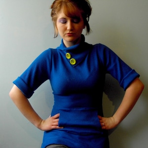 May include: A vibrant blue sweater with a unique design. The sweater features a high collar with two bright yellow buttons, a front pocket, and rolled-up sleeves. The ribbed knit fabric adds texture to the garment.