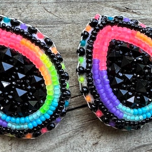 Puede incluir: Dos pendientes de cuentas con un diseño de arcoíris. Los pendientes tienen un centro negro y están rodeados por un arcoíris de colores, incluyendo rosa, naranja, amarillo, verde, azul y morado. Los bordes están bordeados con cuentas negras.