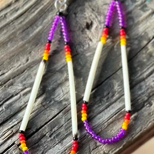 May include: A pair of long, beaded earrings with a white bone center and purple, orange, yellow, and black beads. The earrings are hanging from silver hooks.