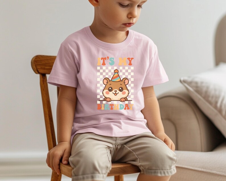 May include: A light pink t-shirt featuring a cartoon hamster wearing a party hat, set against a checkered background. The text "IT'S MY BIRTHDAY" is displayed above and below the hamster. The shirt is worn by a child.