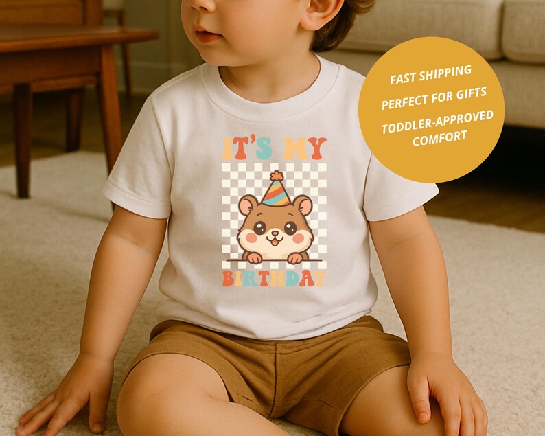 May include: A white toddler t-shirt featuring a cartoon hamster wearing a party hat. The text "IT'S MY BIRTHDAY" is printed above and below the hamster in colorful, retro-style lettering. The background is a checkered pattern.