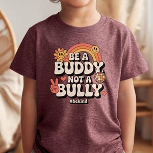 May include: A maroon t-shirt with the words "BE A BUDDY NOT A BULLY" in a retro font, with a rainbow, smiley face, peace sign, and disco ball graphic. The hashtag #bekind is below the text.