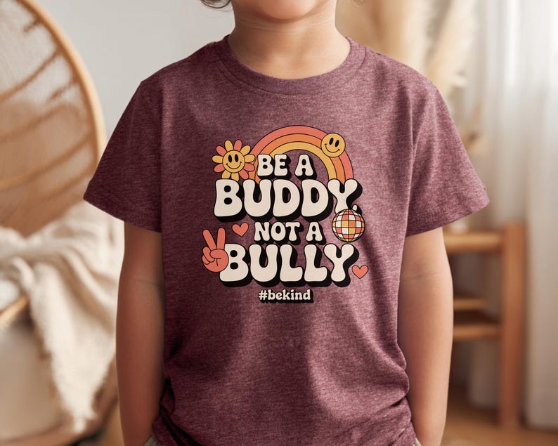 May include: A maroon t-shirt with the words "BE A BUDDY NOT A BULLY" in a retro font, with a rainbow, smiley face, peace sign, and disco ball graphic. The hashtag #bekind is below the text.
