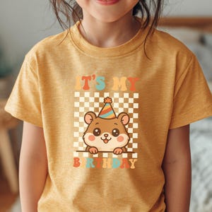May include: Mustard yellow t-shirt featuring a cartoon hamster wearing a party hat. The shirt has the text "IT'S MY BIRTHDAY" in colorful retro lettering, set against a checkered background. A cute and cheerful design for a birthday celebration.