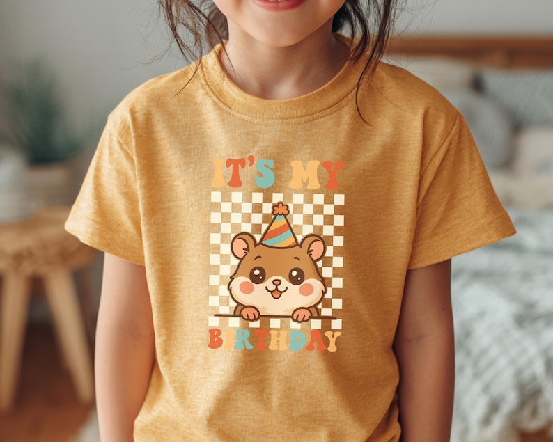 May include: Mustard yellow t-shirt featuring a cartoon hamster wearing a party hat. The shirt has the text "IT'S MY BIRTHDAY" in colorful retro lettering, set against a checkered background. A cute and cheerful design for a birthday celebration.