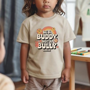 May include: A tan t-shirt with the text "BE A BUDDY NOT A BULLY" in a retro font, featuring a rainbow, flower, peace sign, disco ball, and heart. The hashtag #bekind is below the text. The shirt is worn by a child.