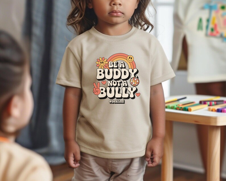 May include: A tan t-shirt with the text "BE A BUDDY NOT A BULLY" in a retro font, featuring a rainbow, flower, peace sign, disco ball, and heart. The hashtag #bekind is below the text. The shirt is worn by a child.
