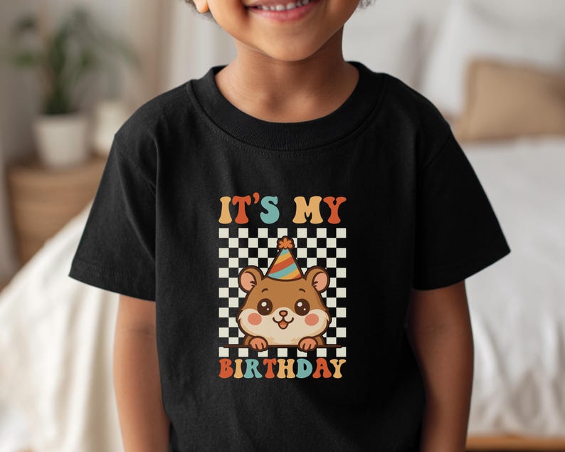 May include: Black t-shirt featuring a cartoon hamster wearing a party hat, set against a checkered background. The text "IT'S MY BIRTHDAY" is displayed in colorful, retro-style lettering. A fun, celebratory design.