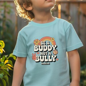 May include: A light blue t-shirt with the text "BE A BUDDY NOT A BULLY" in a retro font, featuring a rainbow, flower, smiley face, peace sign, disco ball, and heart graphics. The hashtag "#bekind" is also included.