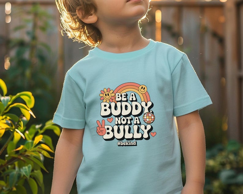 May include: A light blue t-shirt with the text "BE A BUDDY NOT A BULLY" in a retro font, featuring a rainbow, flower, smiley face, peace sign, disco ball, and heart graphics. The hashtag "#bekind" is also included.
