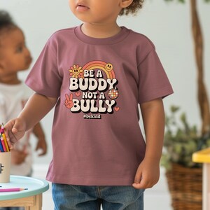 May include: A mauve-colored t-shirt with the text "BE A BUDDY NOT A BULLY" in a retro font, with a rainbow, flower, peace sign, and disco ball graphic. The hashtag #bekind is below the text. The shirt is worn by a child.