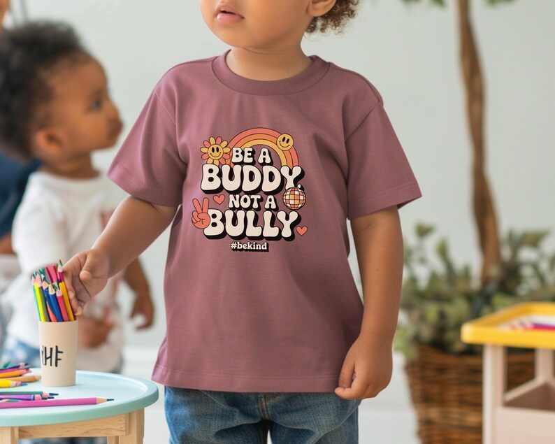May include: A mauve-colored t-shirt with the text "BE A BUDDY NOT A BULLY" in a retro font, with a rainbow, flower, peace sign, and disco ball graphic. The hashtag #bekind is below the text. The shirt is worn by a child.