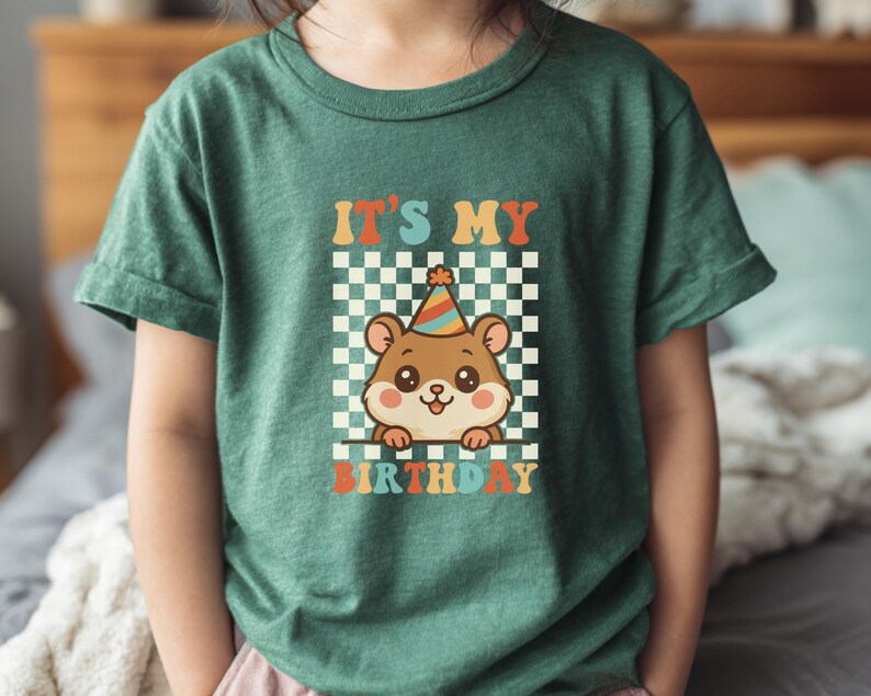 May include: Green t-shirt with the text "IT'S MY BIRTHDAY" in orange, teal, and yellow. A cartoon hamster wearing a party hat is in the center, set against a checkered background. The shirt is made of a soft material.