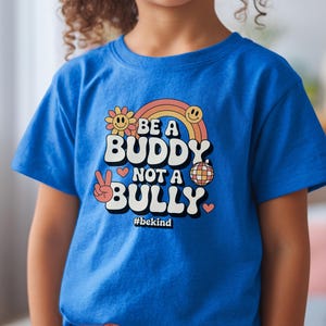 May include: A blue t-shirt with the text "BE A BUDDY NOT A BULLY" in a retro font, with a rainbow, smiley faces, and peace sign graphic. The hashtag #bekind is also included.
