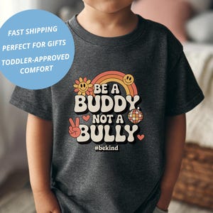 May include: A dark gray t-shirt featuring the text "BE A BUDDY NOT A BULLY" in a retro font, with a peace sign, smiley faces, and a rainbow design. The shirt also includes the hashtag #bekind.