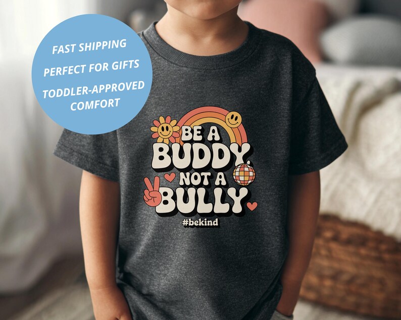 May include: A dark gray t-shirt featuring the text "BE A BUDDY NOT A BULLY" in a retro font, with a peace sign, smiley faces, and a rainbow design. The shirt also includes the hashtag #bekind.
