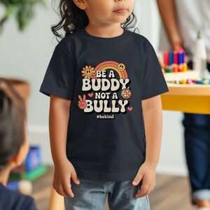 May include: A navy blue t-shirt with the text "BE A BUDDY NOT A BULLY" in a retro design. The design includes a rainbow, flower, peace sign, and disco ball. The hashtag #bekind is also included.