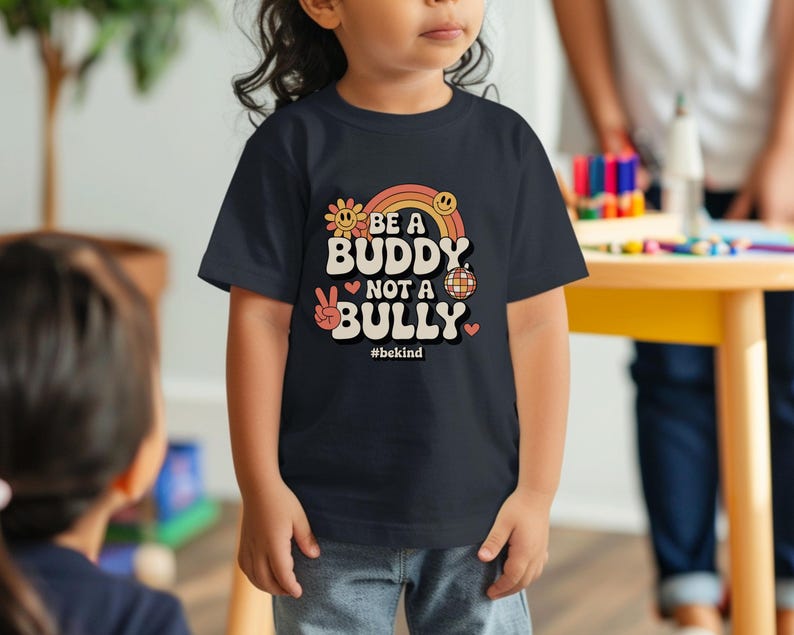 May include: A navy blue t-shirt with the text "BE A BUDDY NOT A BULLY" in a retro design. The design includes a rainbow, flower, peace sign, and disco ball. The hashtag #bekind is also included.