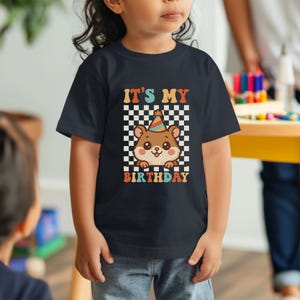 May include: A navy blue t-shirt featuring a cartoon hamster wearing a party hat. The shirt has the text "IT'S MY BIRTHDAY" in a retro font, set against a checkered background. The design is in shades of brown, orange, and white.