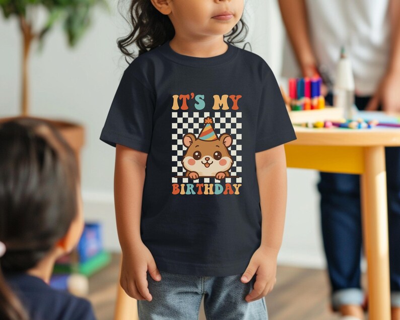 May include: A navy blue t-shirt featuring a cartoon hamster wearing a party hat. The shirt has the text "IT'S MY BIRTHDAY" in a retro font, set against a checkered background. The design is in shades of brown, orange, and white.