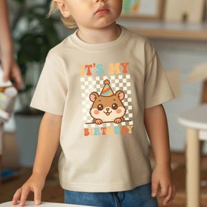 May include: Beige short-sleeved t-shirt featuring a cartoon hamster wearing a party hat. The shirt has the text "IT'S MY BIRTHDAY" in colorful letters, set against a checkered background. The design is centered on the front of the shirt.