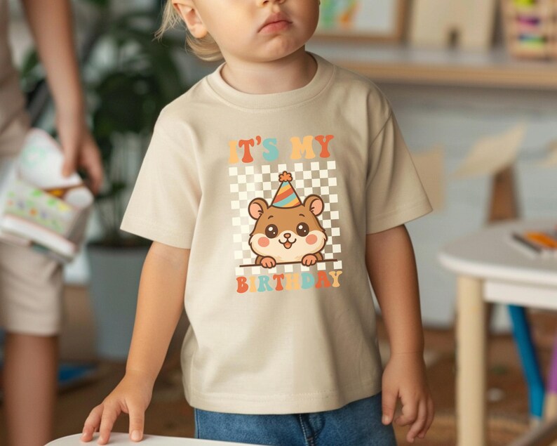 May include: Beige short-sleeved t-shirt featuring a cartoon hamster wearing a party hat. The shirt has the text "IT'S MY BIRTHDAY" in colorful letters, set against a checkered background. The design is centered on the front of the shirt.
