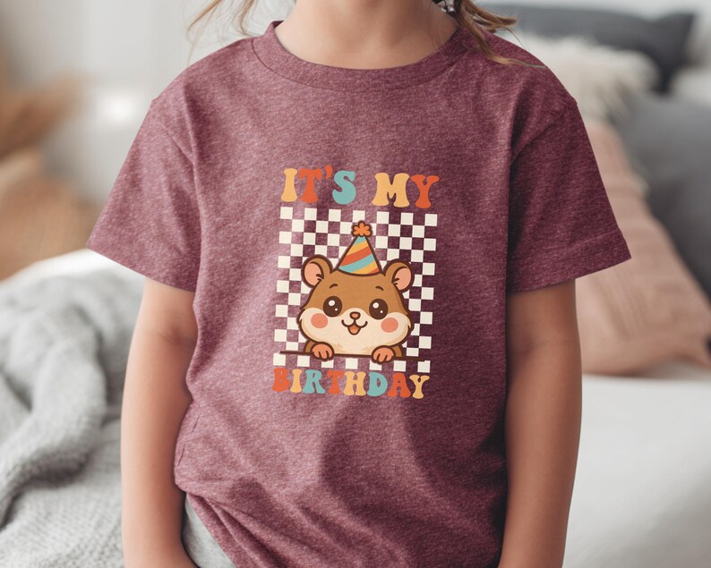 May include: Burgundy t-shirt featuring a cartoon hamster wearing a party hat, set against a checkered background. The text "IT'S MY BIRTHDAY" is displayed in colorful, retro-style lettering. A fun, festive design for a birthday celebration.