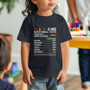 May include: A navy blue t-shirt with the text "BLACK KING NUTRITIONAL FACTS" and a list of values including Melanin, Confidence, Strength, Loyalty, Courage, and Foolishness. The shirt is worn by a child.