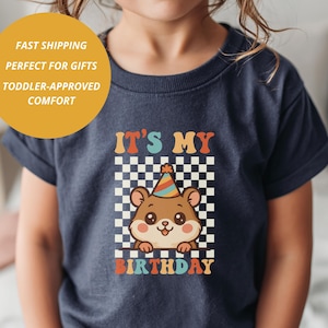 May include: Navy blue toddler t-shirt with a colorful graphic. The graphic features a cute cartoon bear wearing a party hat, set against a checkered background, with the text "IT'S MY BIRTHDAY".