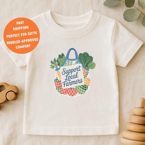 Könnte beinhalten: Cremefarbenes T-Shirt für Kleinkinder mit einer Grafik einer Einkaufstasche voller Gemüse und dem Text "Support Local Farmers". Ein Holzspielzeugauto und ein Holzsteckspielzeug liegen neben dem Shirt.