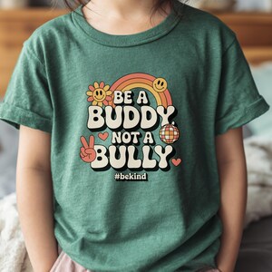 May include: Green t-shirt with the text "BE A BUDDY NOT A BULLY" in a retro font, featuring a rainbow, flower, disco ball, and peace sign graphic. The hashtag #bekind is also included.