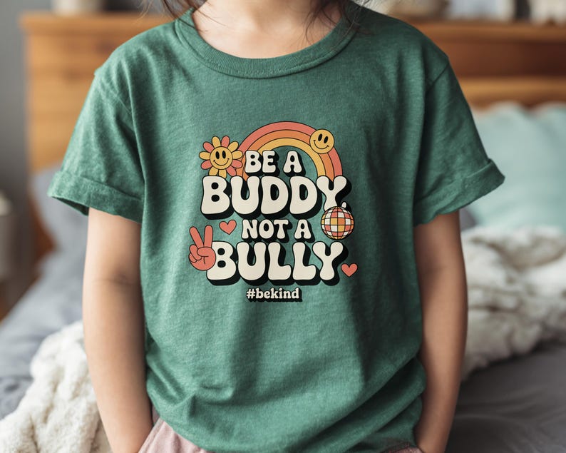 May include: Green t-shirt with the text "BE A BUDDY NOT A BULLY" in a retro font, featuring a rainbow, flower, disco ball, and peace sign graphic. The hashtag #bekind is also included.