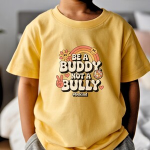 May include: A yellow t-shirt with the text "BE A BUDDY NOT A BULLY" in a retro font, featuring a rainbow, smiley face, flower, disco ball, and peace sign. The hashtag #bekind is also included.